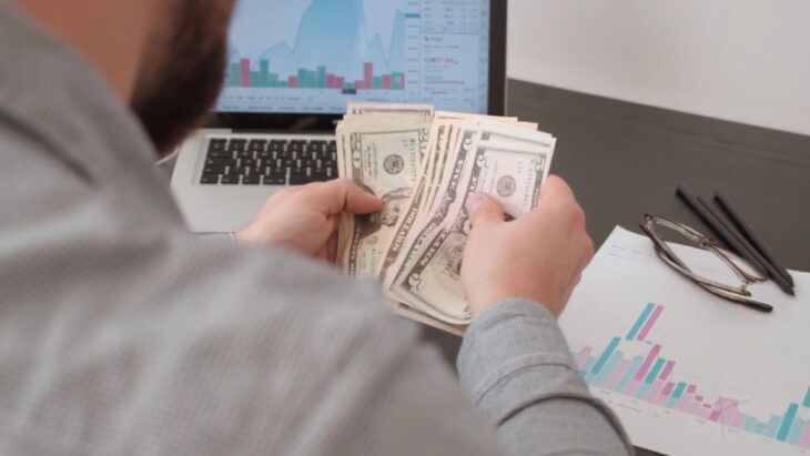 Investor counting cash with stock charts on screen, weighing 2025 portfolio options