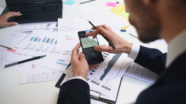 A man reviews stock charts and financial data on his phone surrounded by market analysis reports