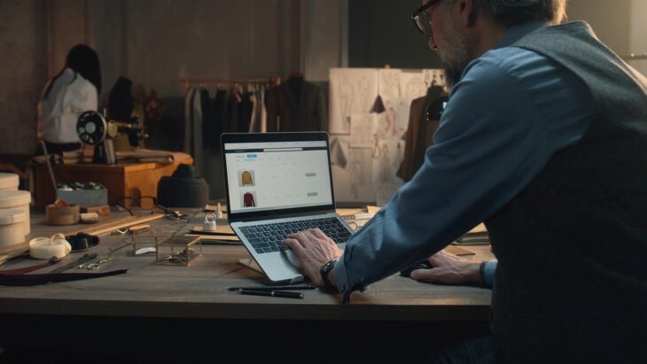 Man crafting detailed product descriptions for clothing items on a laptop in a tailor’s workshop