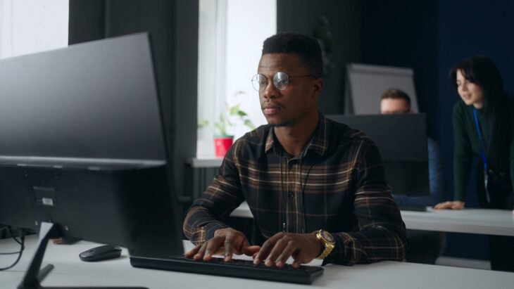 A man working at a computer analyzing risk mitigation strategies with team support in the background