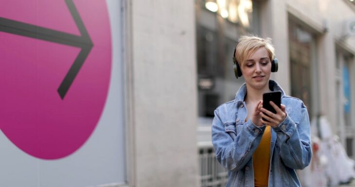 Young woman with headphones looking at her smartphone while walking past a bright arrow sign