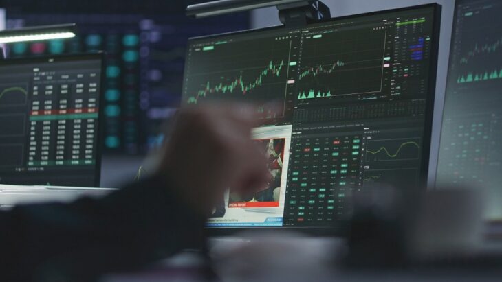Multiple computer monitors displaying stock market charts, price movements, and financial data, with a trader’s hand pointing toward the screen