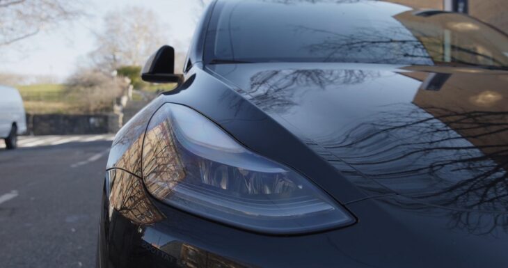 Close-up view of a black Tesla headlight with tree reflections on the hood