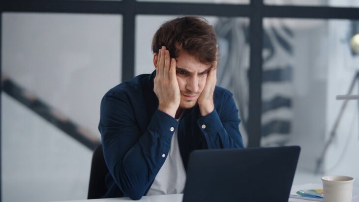 Frustrated young man holding his head while looking at laptop, showing how control over time reduces burnout