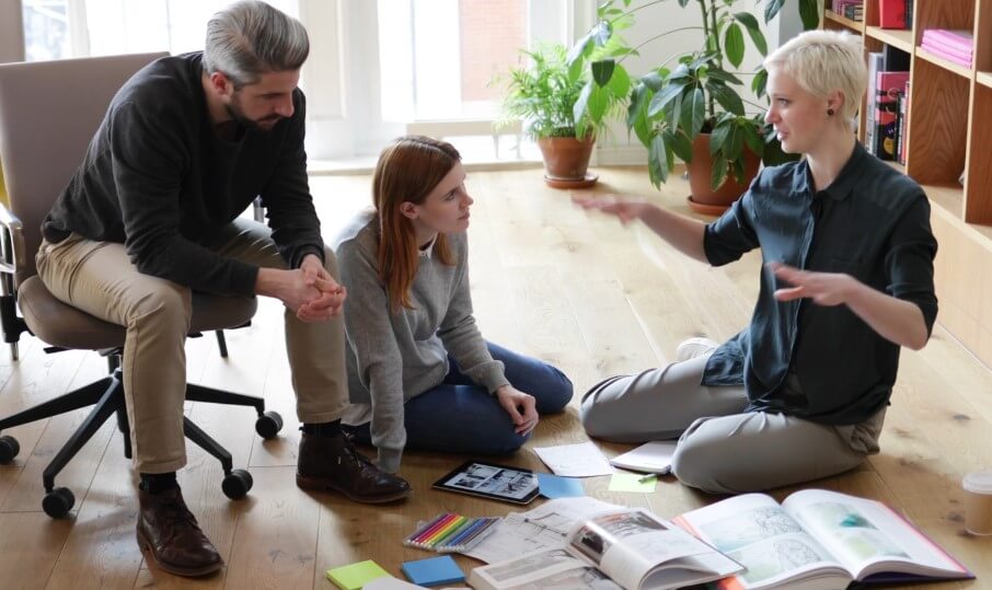 Three people sitting on the floor, surrounded by books and papers, engaged in discussion or study