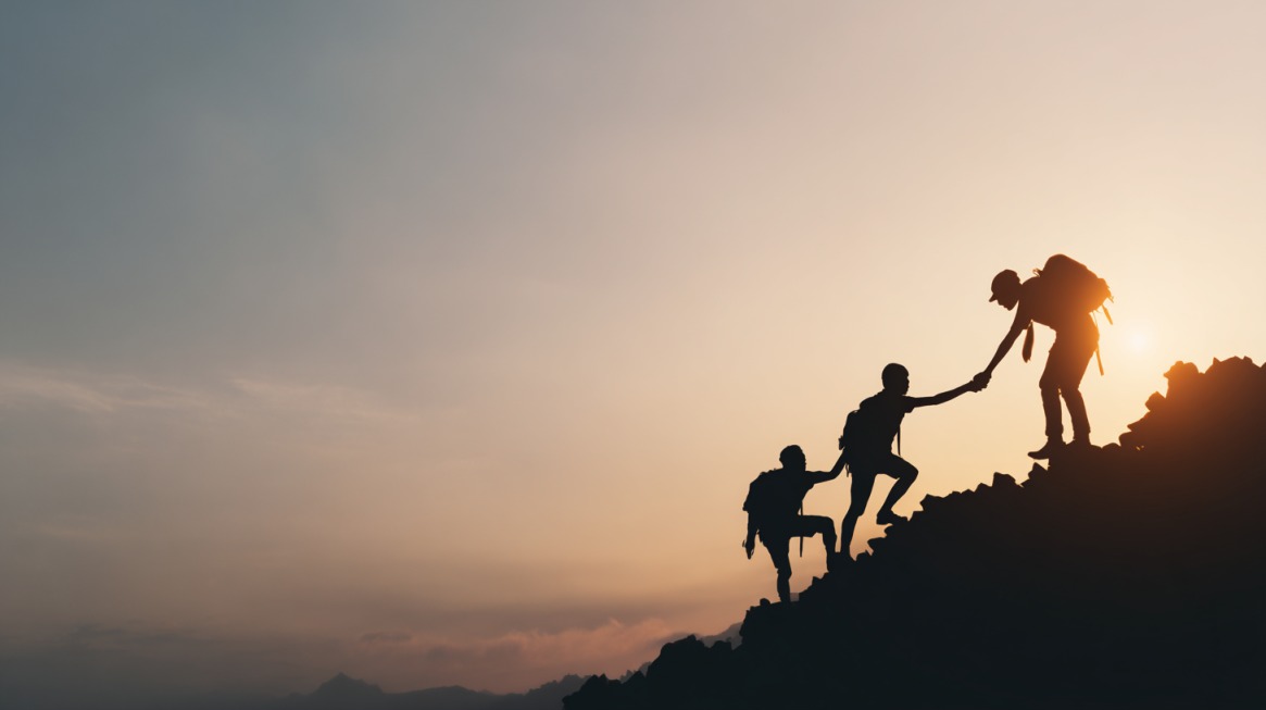 Silhouette of hikers assisting one another while climbing a mountain at sunset