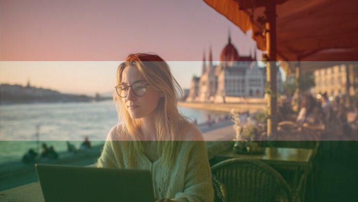 Female freelancer at a Budapest café, laptop open, Parliament dome and Danube softly behind her