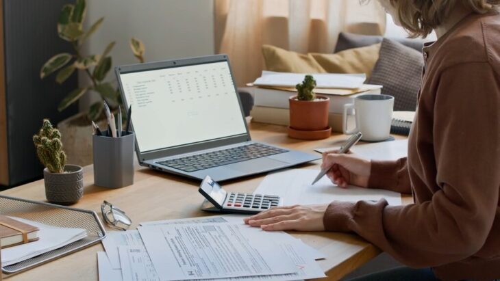 A woman works from home at a desk with a laptop, calculator, and documents, symbolizing the integration of accounting and project management