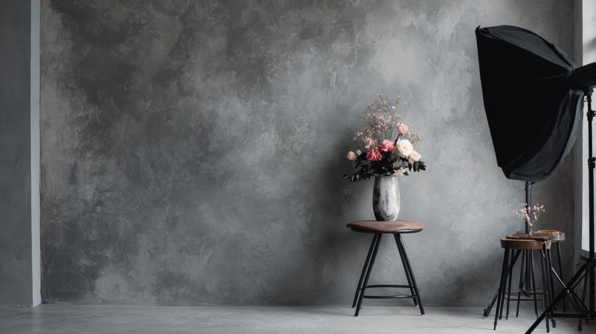 Photography setup with a gray textured wall, softbox lighting, and a floral arrangement in a vase on a stool