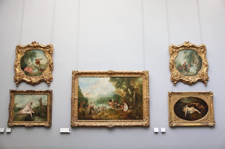 A museum wall displaying five framed classical paintings with ornate golden frames, featuring scenes of people and landscapes