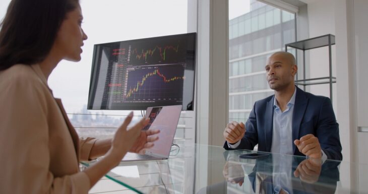 Two business professionals in a modern office discussing investment charts displayed on a large monitor and a laptop
