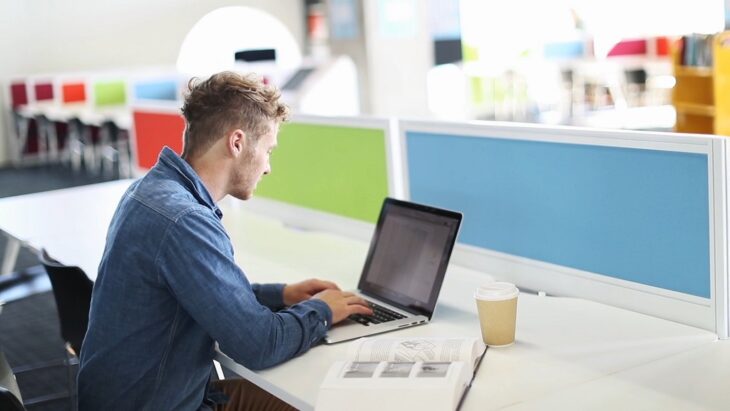 Young designer in a modern coworking space typing on a laptop with coffee and notebook, building layouts for a web design client