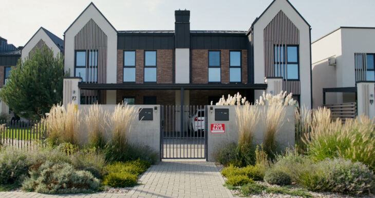 A modern two-story duplex with a “For Sale” sign on the front gate, surrounded by landscaped greenery