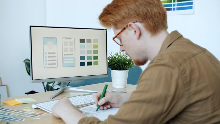 Web designer sketching mobile UI layouts in front of a large monitor displaying color swatches and interface mockups in a minimalist studio