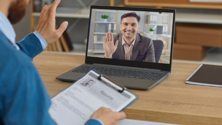 A candidate waves on a laptop screen during a video call as an employer reviews a printed resume