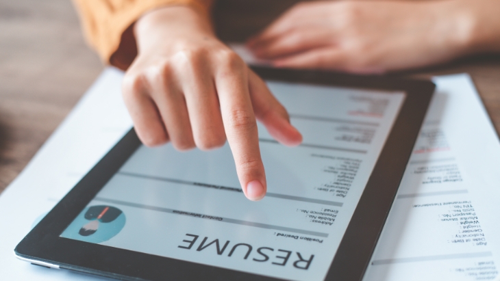 A hand scrolls through a digital resume on a tablet placed over a printed CV