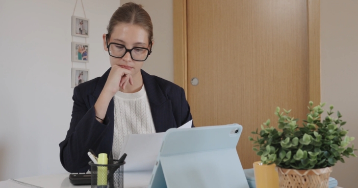 A woman reviews paperwork at her desk with a tablet open beside her