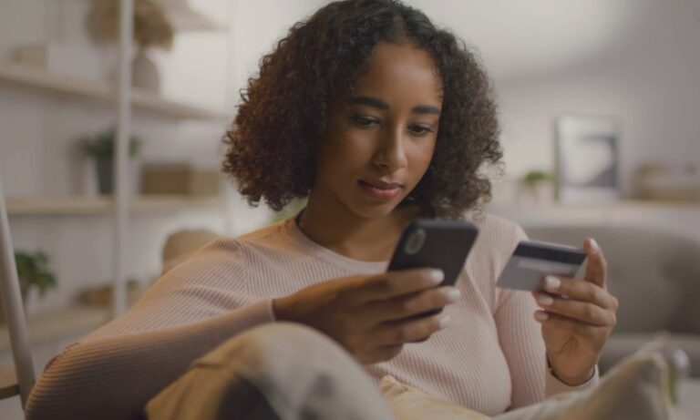 A woman with curly hair is focused on her smartphone while holding a credit card