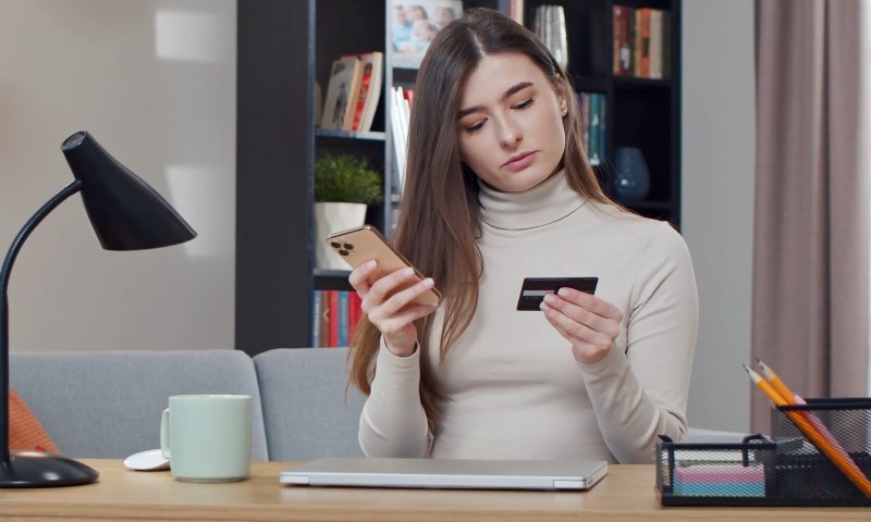 A woman in a beige turtleneck sits at a desk, holding a smartphone and a credit card