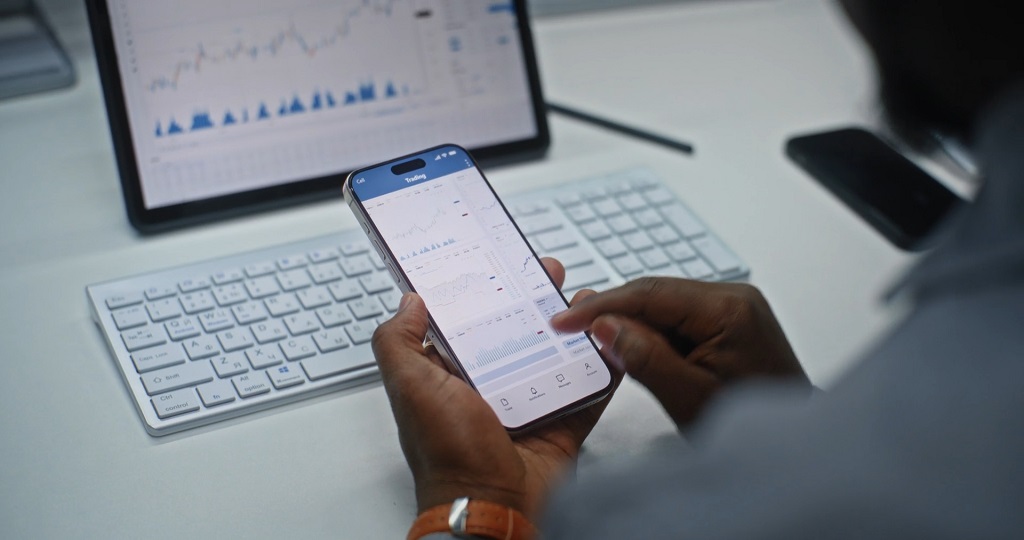 Investor monitoring stock market charts on a mobile trading app while working on a computer