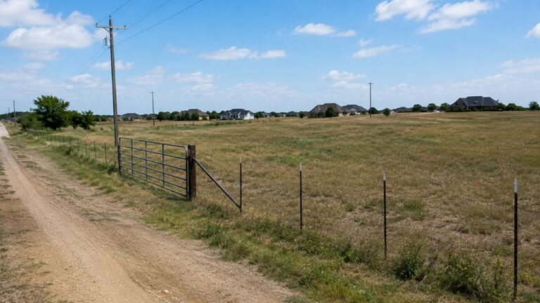 Vacant rural land parcel with dirt road, fencing, utility poles, and residential development in the distance.