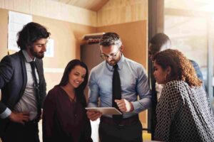 How Many Employees Can One Manager Realistically Supervise - Five business professionals gathered around a tablet, discussing information during a team meeting.