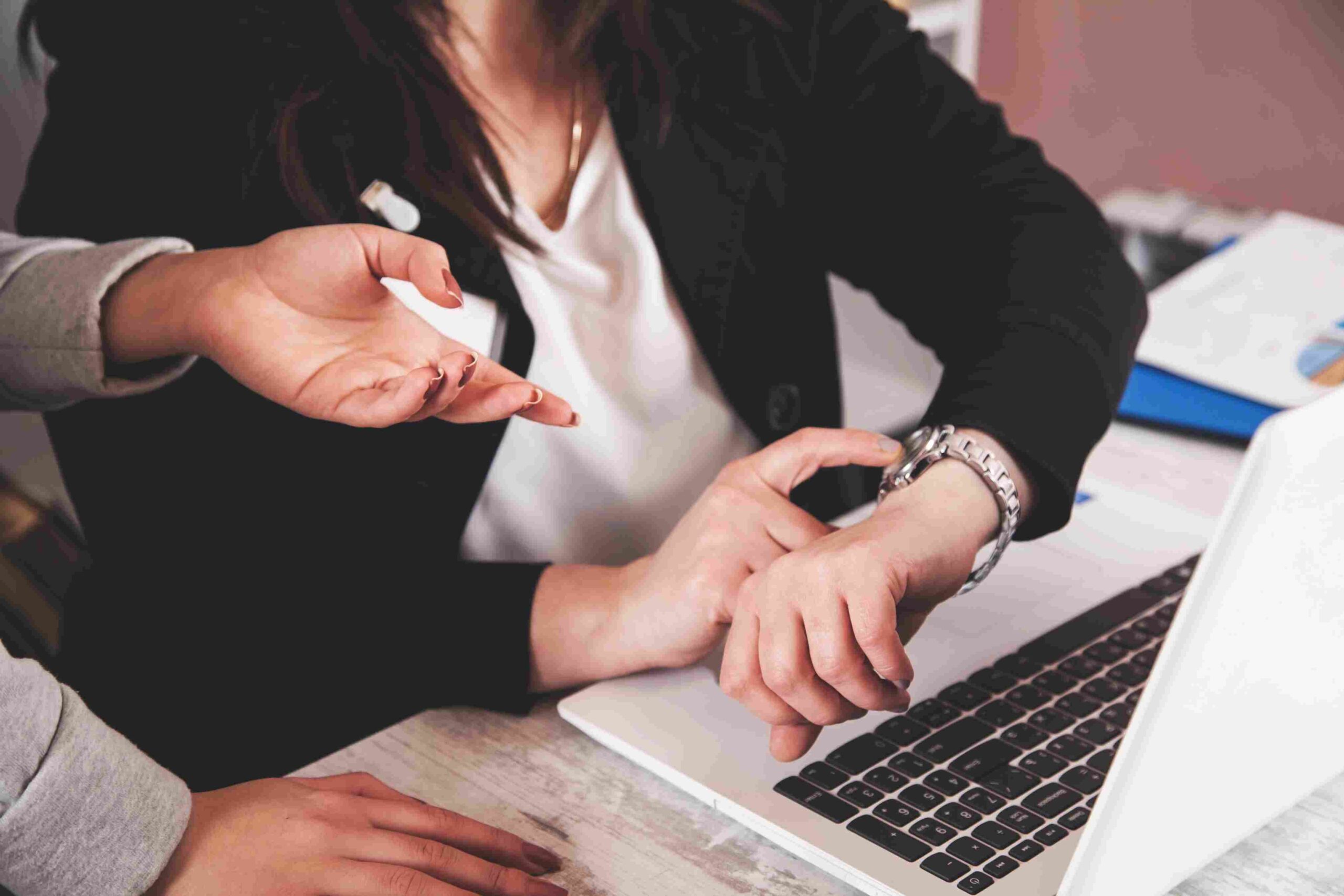 Businesswoman pointing to her wristwatch during a workplace discussion beside a laptop.
