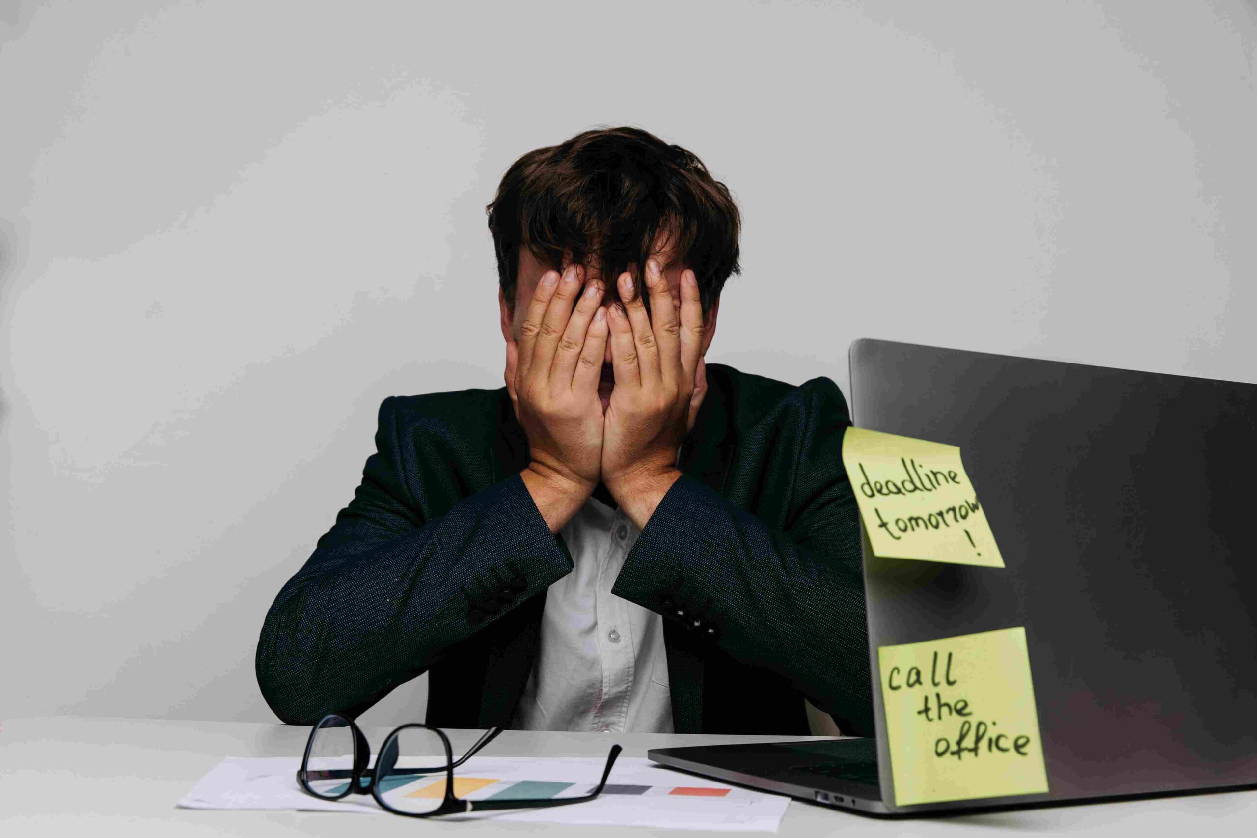Stressed male manager covering his face at a desk with a laptop, paperwork, and deadline reminders.